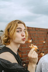 Porträt eines jungen blonden Mannes auf einem Hausdach, der seine Lippen auf die Pizza schürzt - MASF31802