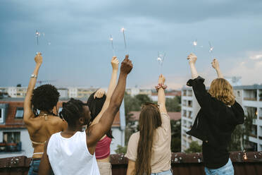 Rückansicht von multirassischen Freunden mit angezündeten Wunderkerzen und erhobenen Händen auf dem Dach - MASF31806