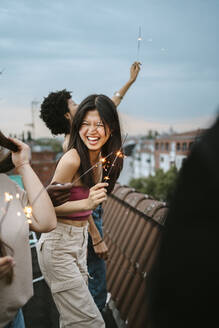 Fröhliche junge Frau, die eine Wunderkerze in der Hand hält und sich mit Freunden auf einem Dach vergnügt - MASF31807