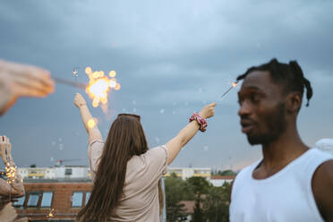 Rear view of woman with arms raised holding lit sparklers against sky - MASF31810