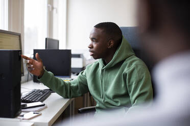 Male trainee in hooded shirt pointing at desktop screen while discussing in office - MASF31830
