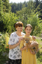Großmutter und Enkelin stehen mit Hund im Garten - LESF00102