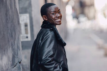 Happy woman wearing leather jacket by wall - GGGF01190