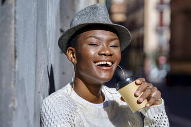 Happy woman holding disposable cup by wall on sunny day - GGGF01199