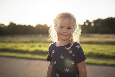 Lächelndes süßes Mädchen mit blonden Haaren steht auf der Straße bei Sonnenuntergang - DWF00594