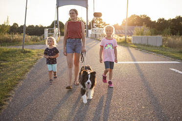 Mädchen und Mutter gehen mit Hund auf der Straße bei Sonnenuntergang - DWF00596