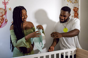 Lächelnde schwangere Frau mit Babykleidung, die bei ihrem Freund zu Hause steht - GGGF01222