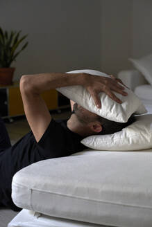 Depressed man covering face with pillow leaning on sofa - VEGF05852