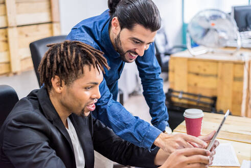 Zwei optimistische multirassische Mitarbeiter in formeller Kleidung surfen mit dem Handy, während sie an einem Tisch in einem modernen hellen Büro mit Dokumenten arbeiten - ADSF36493
