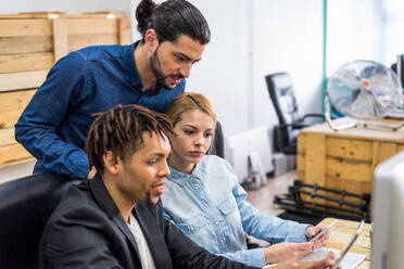 Gruppe optimistischer multirassischer Mitarbeiter in formeller Kleidung, die auf ihrem Handy surfen, während sie an einem Tisch in einem modernen hellen Büro mit Dokumenten arbeiten - ADSF36494