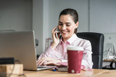 Optimistic female worker talking on smartphone and typing on netbook while sitting at table with takeaway coffee in modern office - ADSF36513