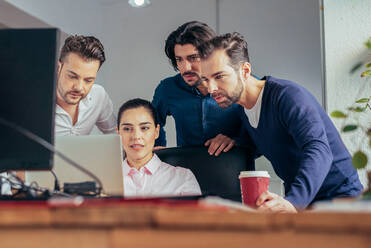 Gruppe lächelnder multirassischer Mitarbeiter in formeller Kleidung, die auf einem Netbook surfen, während sie gemeinsam an einem Geschäftsprojekt in einem modernen hellen Büro arbeiten - ADSF36514