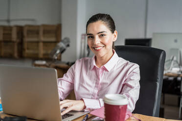 Positiver weiblicher Angestellter in formeller Kleidung mit einer Tasse leckerem Kaffee zum Mitnehmen, der am Tisch in einem hellen, modernen Büro auf ein Netbook schaut - ADSF36515