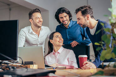 Gruppe lächelnder multirassischer Mitarbeiter in formeller Kleidung, die auf einem Netbook surfen, während sie gemeinsam an einem Geschäftsprojekt in einem modernen hellen Büro arbeiten - ADSF36517