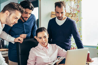 Gruppe lächelnder multirassischer Mitarbeiter in formeller Kleidung, die auf einem Netbook surfen, während sie gemeinsam an einem Geschäftsprojekt in einem modernen hellen Büro arbeiten - ADSF36519