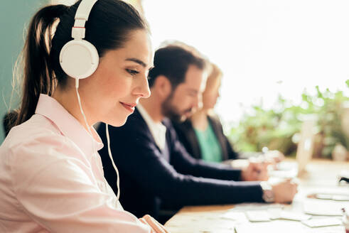 Side view of content female worker listening to audio in headphones while working in light office near colleagues on blurred background - ADSF36522