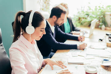 Seitenansicht eines zufriedenen weiblichen Arbeiters, der mit Kopfhörern Audio hört, während er in einem hellen Büro in der Nähe von Kollegen auf unscharfem Hintergrund arbeitet - ADSF36525