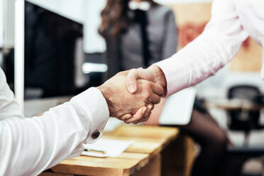 Crop anonymous workers in formal wear shaking hands near wooden table with computer while working in light modern business office - ADSF36536