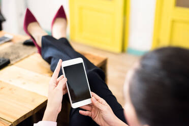 Rückenansicht eines anonymen weiblichen Arbeiters, der auf einem Handy mit schwarzem Bildschirm surft, während er mit den Beinen eines Holztisches in einem modernen Büro sitzt - ADSF36538