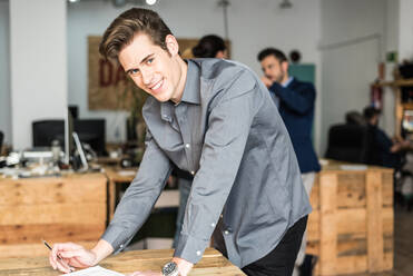 Fokussierter männlicher Arbeiter in formeller Kleidung, der sich Notizen in Dokumenten auf dem Tisch macht, während er in einem hellen modernen Büro arbeitet und in die Kamera schaut - ADSF36554