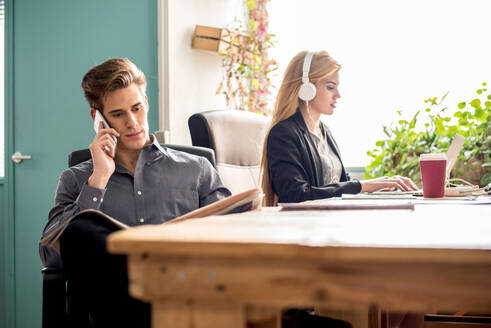 Konzentrierter männlicher Arbeiter, der telefoniert, während er am Tisch neben einer Frau, die auf einem Laptop tippt, Zeitung liest, in einem hellen, modernen Büro - ADSF36555