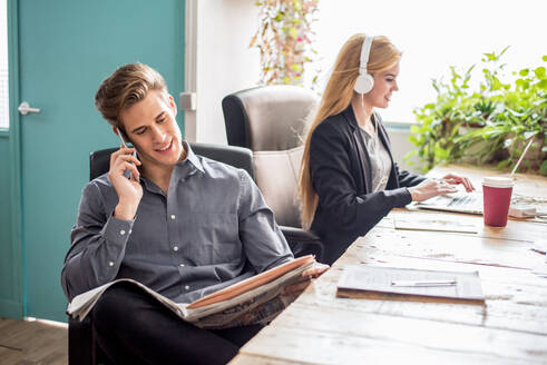 Konzentrierter männlicher Arbeiter, der telefoniert, während er am Tisch neben einer Frau, die auf einem Laptop tippt, Zeitung liest, in einem hellen, modernen Büro - ADSF36556