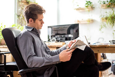 Side view of concentrated male worker in formal wear reading newspaper while sitting near wooden table in light modern office - ADSF36557