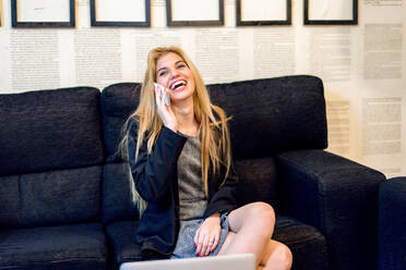 Fröhliche Arbeitnehmerin mit blondem Haar in elegantem Outfit führt ein Telefongespräch, während sie auf einer Couch in einem modernen, hellen Büro sitzt - ADSF36559