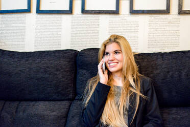 Fröhliche Arbeitnehmerin mit blondem Haar in elegantem Outfit führt ein Telefongespräch, während sie auf einer Couch in einem modernen, hellen Büro sitzt - ADSF36561