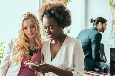 Multirassische schwarze Frau mit weiblicher Kollegin in stilvoller Kleidung surft auf einem Tablet, während sie gemeinsam an einem Geschäftsprojekt in einem modernen hellen Büro arbeiten - ADSF36568