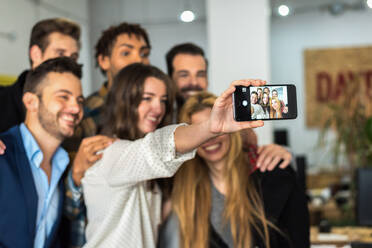 Group of smiling multiracial coworkers in stylish wear taking self portrait on smartphone while standing together in modern light office - ADSF36583