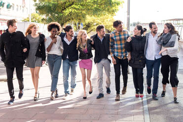 Ganzkörpergruppe von zufriedenen multiethnischen Kollegen in eleganten Outfits, die sich beim Spazierengehen auf einem gepflasterten Gehweg in der Stadt ansehen - ADSF36585