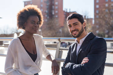 Positive multirassische Mitarbeiter in eleganten Outfits schauen lächelnd in die Kamera, während sie auf einer sonnigen Straße vor Gebäuden in der Stadt stehen - ADSF36589