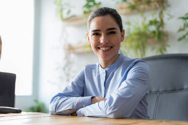 Fröhliche kaukasische Managerin in weißer Bluse mit lockigem Haar, die lächelt und in die Kamera schaut, während sie hinter einem Tisch in einem sonnenbeschienenen Büro sitzt - ADSF36605