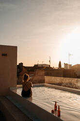 Back view of unrecognizable young female traveler with long hair in stylish black swimsuit sitting in swimming pool and admiring sunset over city during summer holidays in Mallorca - ADSF36613