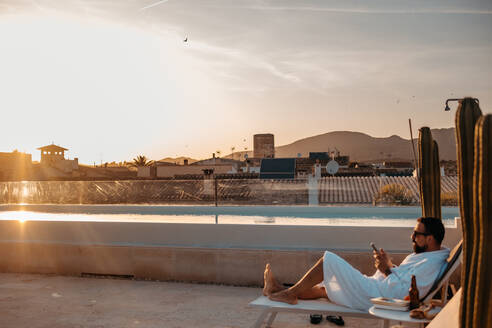 Seitenansicht eines jungen bärtigen männlichen Reisenden in weißem Gewand und Sonnenbrille, der sich auf einer Dachterrasse auf einer Liege entspannt und mit seinem Smartphone gegen den Sonnenuntergang anklickt, während eines Urlaubs auf Mallorca - ADSF36614