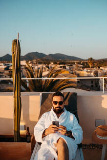 Vorderansicht eines jungen bärtigen männlichen Reisenden in weißem Gewand und Sonnenbrille, der sich auf einer Dachterrasse auf einer Liege entspannt und mit seinem Smartphone Nachrichten gegen den Himmel bei Sonnenuntergang schreibt - ADSF36615