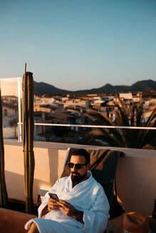 Vorderansicht eines jungen bärtigen männlichen Reisenden in weißem Gewand und Sonnenbrille, der sich auf einer Dachterrasse auf einer Liege entspannt und mit seinem Smartphone Nachrichten gegen den Himmel bei Sonnenuntergang schreibt - ADSF36616