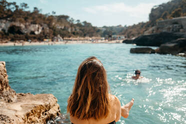 Rückenansicht einer nicht erkennbaren jungen Reisenden mit langen gewellten Haaren in Badekleidung, die sich am felsigen Meeresufer entspannt und die Natur während des Sommerurlaubs auf Mallorca bewundert - ADSF36621