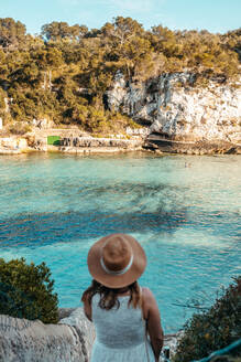 Rückenansicht einer nicht erkennbaren jungen Frau mit Hut und langen, gewellten Haaren, die am felsigen Meeresufer steht und die Natur während der Sommerferien auf Mallorca bewundert - ADSF36625