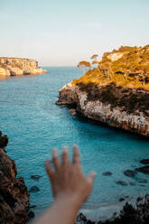 Unbekannter Tourist streckt die Hand aus, während er auf einer felsigen Klippe über dem malerischen Meer mit plätscherndem türkisfarbenem Wasser unter blauem Himmel an einem sonnigen Tag in Mallorca steht - ADSF36630