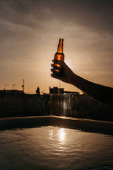 Crop unerkennbar Person demonstriert Flasche Bier beim Chillen im Freibad auf dem Dach gegen bewölkten Sonnenuntergang Himmel in Spanien - ADSF36641