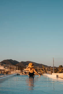 Rückenansicht einer nicht erkennbaren jungen Touristin in einem eleganten schwarzen Badeanzug und einem Strohhut, die im Swimmingpool auf einem Dach steht und die Stadt gegen den wolkenlosen blauen Himmel auf Mallorca bewundert - ADSF36642