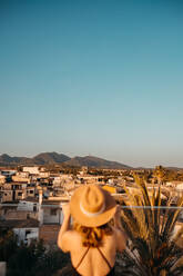 Rückenansicht einer nicht erkennbaren jungen Touristin in modischem schwarzen Badeanzug und Strohhut, die auf einem Dach steht und die Stadt gegen den wolkenlosen blauen Himmel auf Mallorca bewundert - ADSF36644