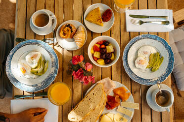 Top view of crop unrecognizable man sitting at wooden table with delicious fried eggs with avocado served on plate near beverages fruits and dessert during breakfast - ADSF36645