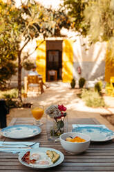 Leckere Spiegeleier mit Speck und Avocado, serviert auf einem Holztisch mit einer Schale frischer Früchte und einem Glas Orangensaft an einem sonnigen Tag im Hinterhof eines Hauses auf Mallorca - ADSF36653