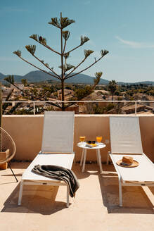 Gläser mit Orangensaft und Sonnenbrille auf einem kleinen Tisch zwischen Sonnenliegen mit Handtuch und Strohhut an einem sonnigen Tag auf einer Terrasse auf Mallorca - ADSF36657
