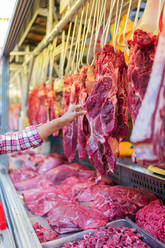 Unrecognizable customer touching and inspecting piece of raw meat hanging over stall while visiting butcher store on market - ADSF36662