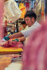 Man in white robe and latex gloves using sharp knife to cut piece of fat from fresh meat during work in butchery - ADSF36664