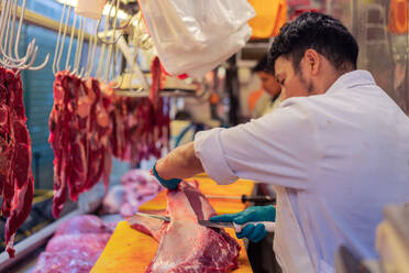 Man in white robe and latex gloves using sharp knife to cut piece of fat from fresh meat during work in butchery - ADSF36665